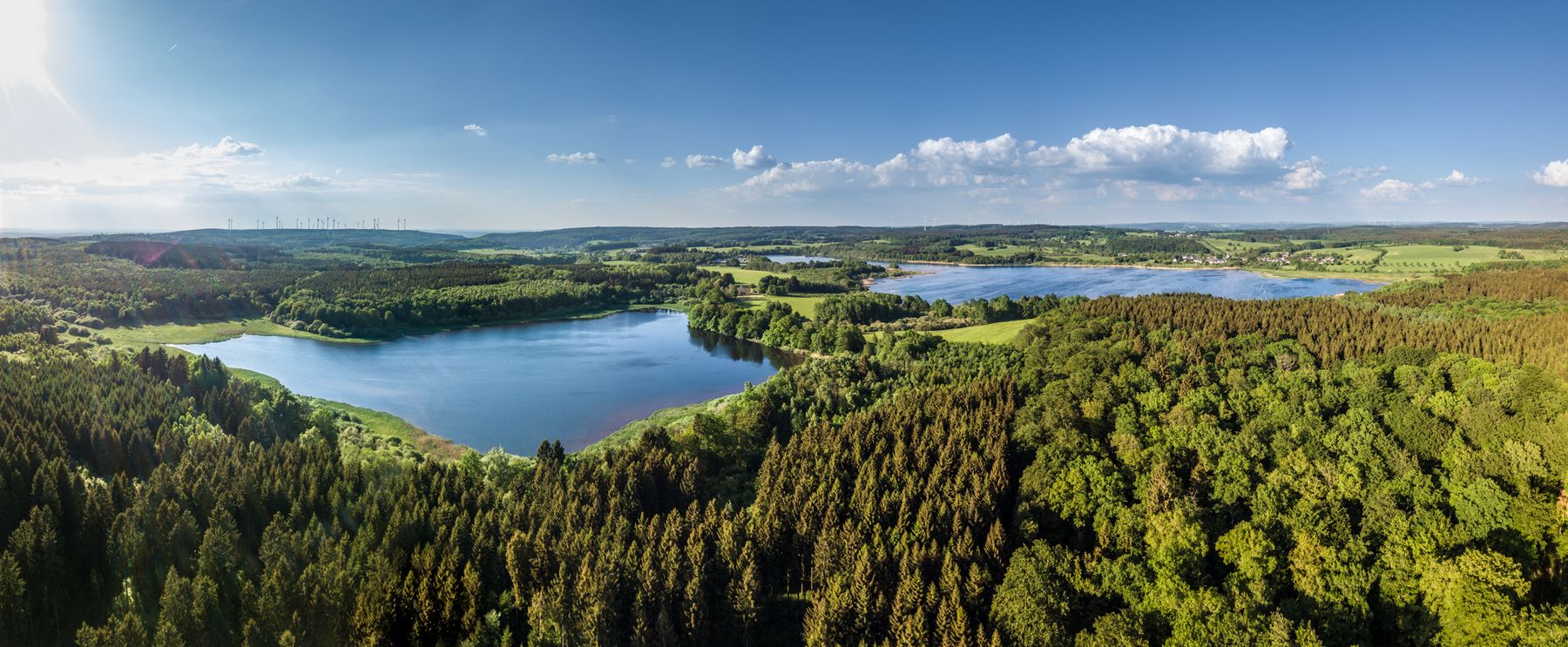 Ein Panoramablick aus der Luft auf einen üppigen grünen Wald, der zwei Seen umgibt, unter einem klaren blauen Himmel mit vereinzelten Wolken und sanften Hügeln im fernen Hintergrund.